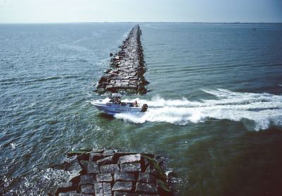Boat speeding along a long, rocky jetty in open water, creating a frothy wake under a clear blue sky.