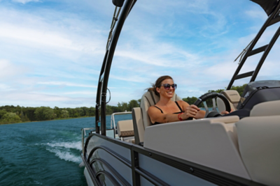 Women steering a Harris Sunliner pontoon boat on a body of water with trees visible in the background. 