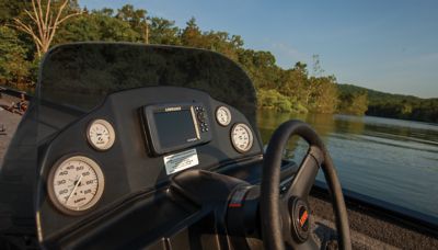 The image shows the dashboard of a Lowe Legacy Console 2022 boat featuring Lowrance technology. The console includes a Lowrance fish finder or navigation device mounted in the center, surrounded by four analog gauges displaying various metrics. The steering wheel is visible on the right side of the console. In the background, there is a serene lake with calm water and lush green trees along the shoreline under a clear sky.