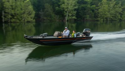 A black Lowe Legacy boat with an orange stripe speeds across a calm lake, creating gentle ripples in the water. The boat is powered by a Mercury outboard motor and carries two people: an adult in a white shirt and cap at the helm, and a child in a yellow life jacket seated beside them. The lake is surrounded by lush green trees.