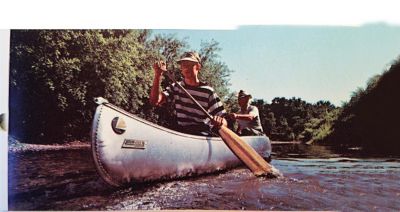 Two people are paddling a canoe on a calm river. The person in the front is wearing a striped shirt and a hat, while the person in the back is also wearing a hat. The surrounding area is lush with greenery, indicating that they are likely in a natural, possibly remote location. The image captures an outdoor recreational activity, highlighting the enjoyment of nature and adventure.