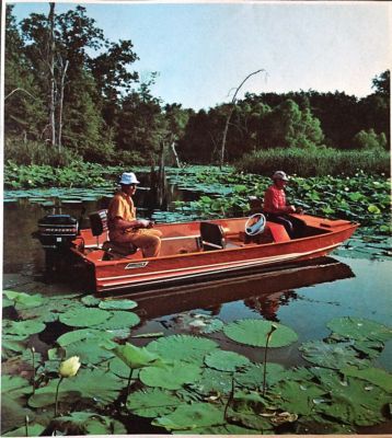 Two people in a small red motorboat on a lake filled with lily pads and surrounded by trees. The boat is equipped with an outboard motor, and the individuals are seated, one at the front and one at the back of the boat. The scene is calm and serene, with lush greenery in the background.