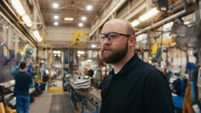A Harris employee stands in a factory, surrounded by various machinery and equipment. The factory environment includes overhead lighting, industrial tools, and other workers engaged in tasks.