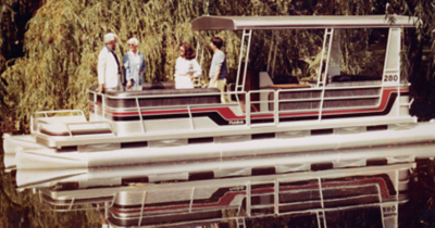 The image shows a historical Harris pontoon boat floating on a calm body of water. The boat has a white and silver color scheme with red and black stripes along the sides. 