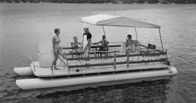 Black and white photograph of a historic Harris pontoon boat on a body of water featuring a family grilling.