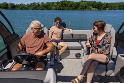 The image shows three people sitting on a Harris pontoon boat. The boat is on a body of water with a forested shoreline in the background. The individuals are seated in the boat's seating area, which includes cushioned seats and a steering wheel visible to the left. The interior of the boat features gray upholstery and flooring, with some storage compartments and speakers visible.