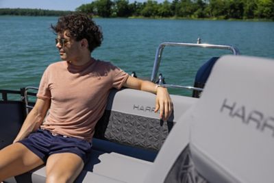 The image shows a person sitting on a Harris pontoon boat. The person is wearing a light brown t-shirt and dark blue shorts, and is seated on the boat's cushioned seating area. The background features a body of water with trees along the shoreline. The word "HARRIS" is visible on the backrest of the seat in front of the person and also on the seat where the person is sitting.