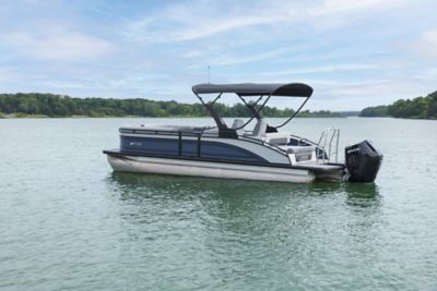 The image shows a pontoon boat floating on a calm body of water with a forested shoreline in the background. The boat has an outboard motor and is equipped with a canopy providing shade over the seating area. The sky is partly cloudy, and the water appears to be tranquil.