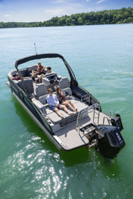 The image shows a motorboat with four people onboard, cruising on a calm, greenish-blue lake. The boat has an open deck with seating areas and is equipped with a large outboard motor labeled "400". The background features a tree-lined shore under a clear blue sky.