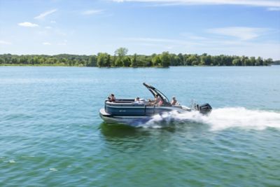 The image shows a pontoon boat cruising on a calm lake with a backdrop of lush green trees and a clear blue sky. The boat is moving at a high speed, creating white water trails behind it. There are several people on the boat enjoying the ride.