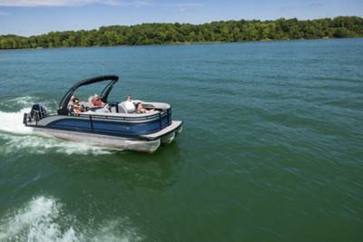 The image shows a pontoon boat with several people on board, cruising on a large body of water. The background features lush green trees and a clear blue sky.