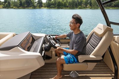 The image shows a person sitting in the driver's seat of a boat, holding the steering wheel. The boat is on a body of water with trees visible in the background. The person is wearing a gray t-shirt and blue shorts with a pattern. The seat has "HARRIS" written on it.