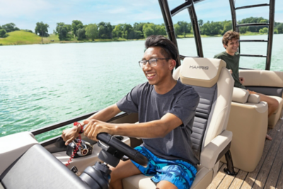 The image shows two people sitting on a boat. The person in the foreground is holding the steering wheel and appears to be driving the boat, while the person in the background is seated and looking out at the water. The boat is on a body of water with trees and greenery visible in the background. The seats are labeled "HARRIS."