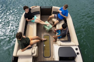 The image shows four people sitting on a boat in the middle of a body of water. The boat has beige cushioned seats and wooden flooring. There is a green cooler in the center, and some of the individuals are holding drinks. The water around the boat is calm, and there are no visible landmasses or other boats nearby.