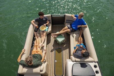 The image shows four individuals relaxing on a boat. The boat is on a body of water, and the individuals are seated on cushioned benches arranged in a U-shape. One person is holding a bowl of snacks, while the others appear to be conversing or resting. The floor of the boat has wooden paneling, and there is a small table in the center with an item placed on it.