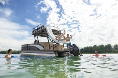 The image shows a pontoon boat on a body of water with several people enjoying recreational activities. Two individuals are captured mid-air as they jump off a slide attached to the boat, while two others are in the water nearby. The sky is partly cloudy, and there are trees in the background along the shoreline.