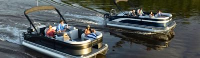 Two Lowe pontoon boats with passengers cruising side-by-side on a calm lake surrounded by trees