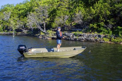 A person stands in a 14 foot boat fishing on a lake with trees and a rocky shore in the background.