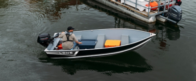 Image of a Lowe WV 1470 utility boat on a body of water near a docked pontoon boat. The boat has a sleek black and silver exterior with the brand name 'LOWE' visible on the side. Inside, there are two bench seats and an orange cooler placed towards the front. A person wearing a cap and beige shirt is seated at the rear, operating the outboard motor attached to the back of the boat.