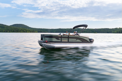 A Lowe SF fishing pontoon boat speeds across a lake with several passengers seated under a secured dark Bimini; water splashes behind the boat as green trees line the shore in the background. 