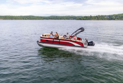 A red-and-white Lowe SS Pontoon Boat speeds across a lake, with several people seated, as the wake trails behind, and a green shoreline with trees in the distance