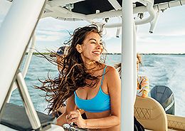 A woman in a blue swimsuit smiles joyfully on a speeding boat, wind in her hair, with a water trail visible in the background.